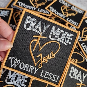 Könnte beinhalten: Ein schwarzer Stoff-Patch mit goldenen Nähten und weißem Text. Der Patch lautet "Pray More Worry Less!" mit einem stilisierten Herz- und Kreuzdesign. Das Herz hat das Wort "Jesus" im Inneren.