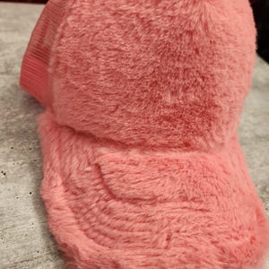 Peut inclure: Casquette de camionneur rose et duveteuse avec un dos en filet. La casquette a une visière incurvée et une texture douce et moelleuse. La couleur est un rose clair vif.