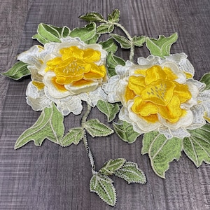 May include: Two yellow embroidered flowers with white centers and green leaves. The flowers are attached to a stem with leaves.