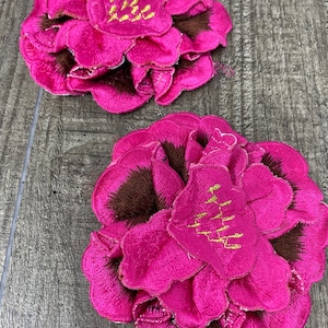 Pode incluir: Dois patches bordados de flores fúcsia com centros castanhos e detalhes de texto dourado. Os patches apresentam pétalas em camadas, criando um efeito tridimensional. Estes patches florais são ideais para adicionar um toque decorativo a roupas ou acessórios.