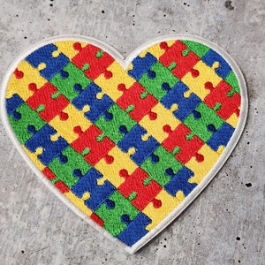 May include: A heart-shaped patch with a white outline, filled with a pattern of colorful puzzle pieces. The puzzle pieces are red, yellow, blue, and green.