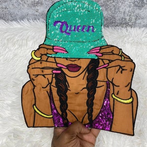 May include: A brown-skinned woman with long braids is wearing a turquoise sequin bucket hat with the word "Queen" embroidered in purple. She is wearing a purple sequin top and has pink nail polish on her nails.