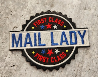以下が含まれることがあります： 黒と白の刺繍パッチで、「First Class」と「Mail Lady」の文字が青と赤の星で書かれています。