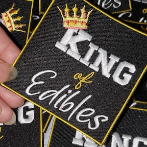 Puede incluir: Un parche negro con ribete dorado y bordado blanco. El parche dice "King of Edibles" con una corona dorada sobre la palabra "King".