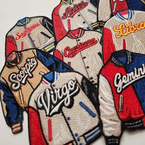 May include: A set of 10 embroidered iron-on patches featuring zodiac signs. The patches are in the style of varsity jackets with red, white, and blue color schemes. The signs include Aries, Taurus, Gemini, Cancer, Leo, Virgo, Libra, Scorpio, Sagittarius, and Pisces.