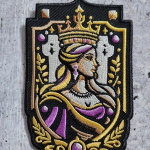 May include: Embroidered patch featuring a crowned woman with a purple and gold robe, set against a black background with gold accents. The patch has a medieval theme.