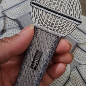 May include: Embroidered microphone patch in shades of gray and black. The microphone head features a detailed grid pattern, with a black horizontal bar. The handle has a rectangular black detail. Patches are visible in the background.