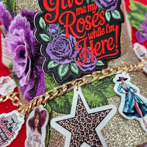 May include: A gold glitter hat with a patch that says "Give me my Roses while I'm Here!" in red script. The hat has purple rose embroidery and a gold chain with charms, including a leopard print star and a woman in a dress.