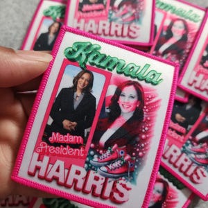 May include: A white fabric patch with pink stitching around the edges. The patch features a colourful image of Kamala Harris with the text "Kamala Harris" and "Madam President" in green and pink lettering.