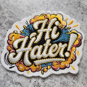 May include: Embroidered patch with the words "Hi Hater!" in white script outlined in gold. The text is set against a black and teal background with yellow and pink accents, all on a white cloud-shaped patch.