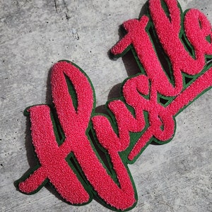 Puede incluir: Parche de tela rosa y esponjosa con la palabra "Hustle" escrita en una fuente en negrita y cursiva. El parche tiene un contorno verde.