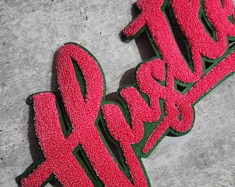 May include: Pink fluffy fabric patch with the word "Hustle" written in a bold, cursive font. The patch has a green outline.