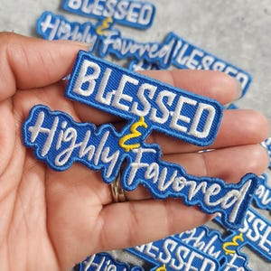 May include: Embroidered patches in blue and white, featuring the phrases "BLESSED" and "Highly Favored". The patches have a blue base with white stitching and a yellow accent. The patches are held in a hand.