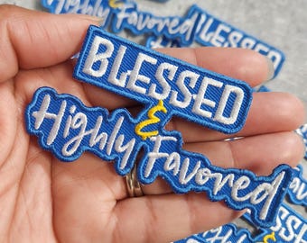 Könnte beinhalten: Blaue und weiße gestickte Aufnäher mit den Worten "BLESSED" und "Highly Favored" in Schreibschrift. Die Aufnäher haben einen blauen Hintergrund mit weißen Nähten und einem gelben Akzent. Die Aufnäher werden in einer Hand gehalten.