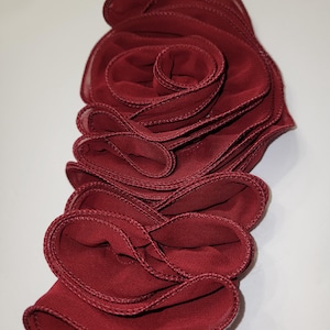 May include: A burgundy fabric flower with layered, ruffled petals. The edges are stitched with a matching thread, creating a textured, three-dimensional effect. The flower is a deep red color and appears to be made of a lightweight material.