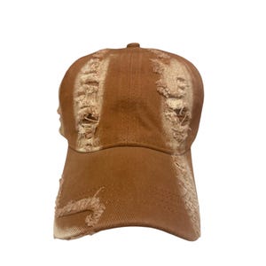 May include: A brown baseball cap featuring a distressed, frayed texture across the front panels and the curved brim. The hat is constructed from a solid-colored fabric with visible stitching details and a button at the top center of the crown.