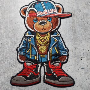 May include: Embroidered patch of a teddy bear wearing a blue denim jacket, jeans, red sneakers, and a blue and red baseball cap with the text "Good Life" on it.