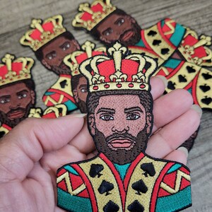 May include: An embroidered patch featuring a king with a red and gold crown, wearing a green, red and gold patterned suit with black spades on the front.