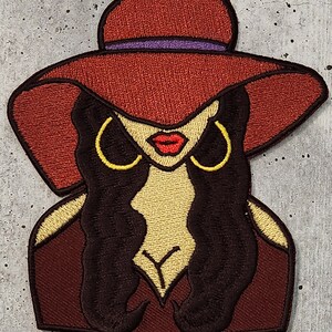 May include: Embroidered patch of a woman with long dark hair wearing a large red hat with a purple band. She has large gold hoop earrings and red lips.