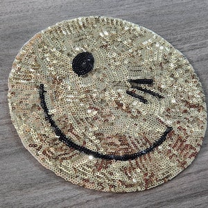 Puede incluir: Parche de cara sonriente de lentejuelas doradas con contorno negro. La cara sonriente está guiñando un ojo.