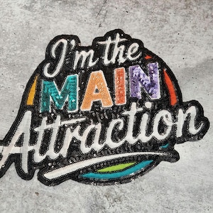 Puede incluir: Un parche negro, blanco y arcoíris de lentejuelas con el texto "I'm the main attraction" en una fuente cursiva.