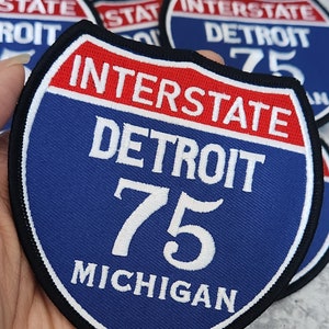 以下が含まれることがあります： 「Interstate Detroit 75 Michigan」と白文字で書かれた青と赤の州間高速道路標識パッチ。
