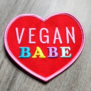Pode incluir: Patch vermelho em forma de coração com costuras cor-de-rosa ao redor das bordas. O patch diz "VEGAN BABE" em letras bordadas coloridas.
