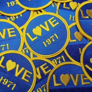 Puede incluir: Primer plano de varios parches bordados azules y dorados con la palabra "LOVE" y el año "1971" en ellos. Los parches son circulares y tienen una forma de corazón en el centro.