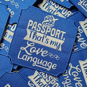 以下が含まれることがあります： 飛行機と地球儀のデザインが施された、白い刺繍で「PASSPORT. That's my Love Language」と書かれた青い布のパッチ