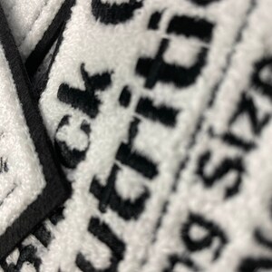 May include: Close-up of white towels with black text. The towels have a textured surface and a black border. The text is partially visible, with the words "Black" and "Buying Size" discernible. The image is well-lit, highlighting the contrast between the black and white.