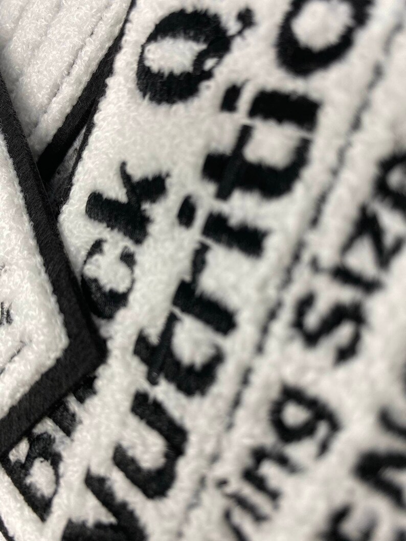 May include: Close-up of white towels with black text. The towels have a textured surface and a black border. The text is partially visible, with the words "Black" and "Buying Size" discernible. The image is well-lit, highlighting the contrast between the black and white.
