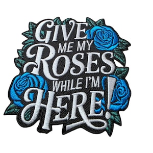 May include: Embroidered patch with the text "GIVE ME MY ROSES WHILE I'M HERE" in white script on a black background. The patch features blue roses and green leaves around the edges, creating a floral design.