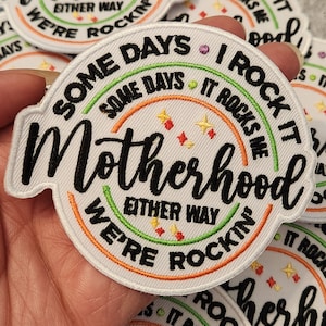 Könnte beinhalten: Gestickter Aufnäher mit dem Wort "Motherhood" in einem kreisförmigen Design. Der Aufnäher enthält die Sätze "Some days, I rock it, it rocks me, either way, we're rockin'". Der Aufnäher hat einen weißen Hintergrund mit schwarzen, orangefarbenen, grünen und gelben Akzenten.