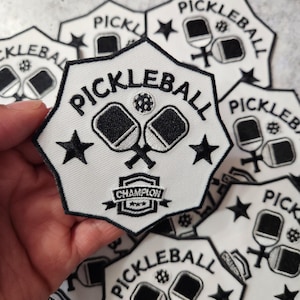 May include: White and black embroidered patch with the word "PICKLEBALL". The patch features two crossed pickleball paddles, a pickleball, and stars. The word "CHAMPION" is also displayed.