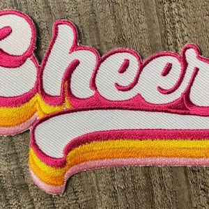 Può includere: Una toppa ricamata bianca con la parola "Cheer" in un font retrò e colorato. Le lettere sono contornate di rosa e giallo.