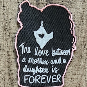 May include: Black embroidered patch with pink stitching shaped like two people hugging. The text reads "The love between a mother and a daughter is FOREVER".