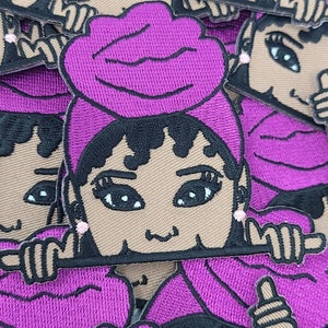 May include: A set of embroidered iron-on patches featuring a cartoon character with brown skin, black hair, and a purple headscarf. The character is smiling and has large eyes. The patches are perfect for adding a touch of personality to clothing, bags, or other accessories.
