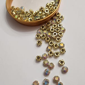 Peut inclure: Gros plan sur des perles en métal doré en forme de fleur avec des strass irisés multicolores. Certaines perles sont dans un récipient doré. Les perles sont dispersées sur une surface blanche. Idéal pour la fabrication de bijoux et l'artisanat.