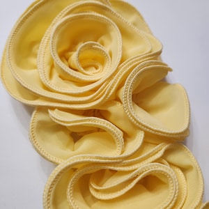 May include: A large, pale yellow fabric flower with multiple layers of petals. The edges of each petal are finished with a delicate, light yellow stitching. The flower is set against a white background.
