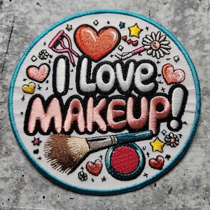 Puede incluir: Parche redondo blanco bordado con un borde turquesa. El parche presenta un texto rosa y blanco que dice "I Love Makeup!" con un corazón rojo, una brocha de maquillaje y un rubor rosa.