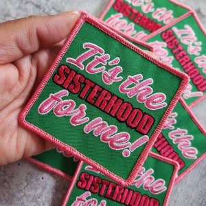 May include: Green fabric patch with pink stitching and the text "It's the Sisterhood for me!" in pink and black lettering.