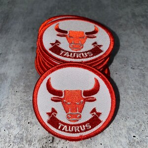 Könnte beinhalten: Ein Stapel roter und weißer gestickter Bügelbilder mit einem Stierkopf und dem Wort "TAURUS" in weißen Buchstaben.