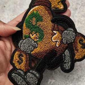May include: An embroidered patch featuring a cartoon character holding a money bag. The character is wearing a brown hat and the money bag has a green dollar sign on it.