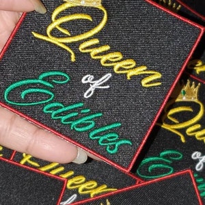 Puede incluir: Un parche de tela negro con borde rojo. El parche presenta una corona dorada con una joya verde, y el texto "Queen of Clubs" en letras doradas y verdes.