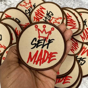Puede incluir: Primer plano de un parche bordado marrón y beige con una corona roja y el texto "SELF MADE" en letras negras y rojas.