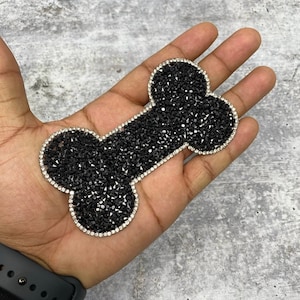 Puede incluir: Parche con forma de hueso de perro de pedrería negra. El parche está bordeado con pedrería transparente.