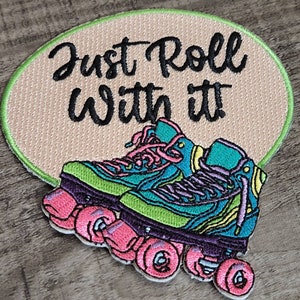 May include: Embroidered patch with teal, green, and purple roller skates and pink wheels. The patch has the text "Just Roll With It" in black script on a tan oval background with a green border.