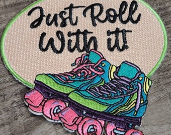 May include: Embroidered patch featuring a pair of teal, green, and purple roller skates with pink wheels. The patch has the text "Just Roll With It" in black script on a tan oval background with a green border.