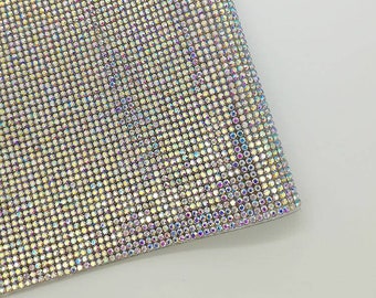 May include: Close-up of a shimmering, iridescent fabric. The material is covered in small, closely-set, reflective rhinestones, creating a dazzling, textured surface. The color shifts with light, displaying a spectrum of colors.
