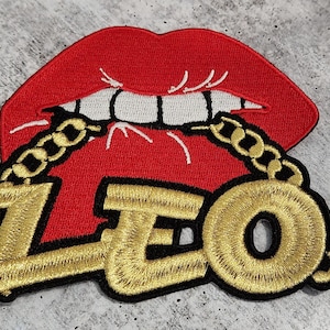 Op de afbeelding: Rode lippen met een gouden ketting en het woord "LEO" in gouden letters. De ketting is om de lippen gewikkeld en het woord "LEO" is aan de ketting bevestigd.